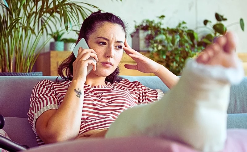 Worried woman with broken leg in plaster cast lying down on sofa at home and talking on the phone.